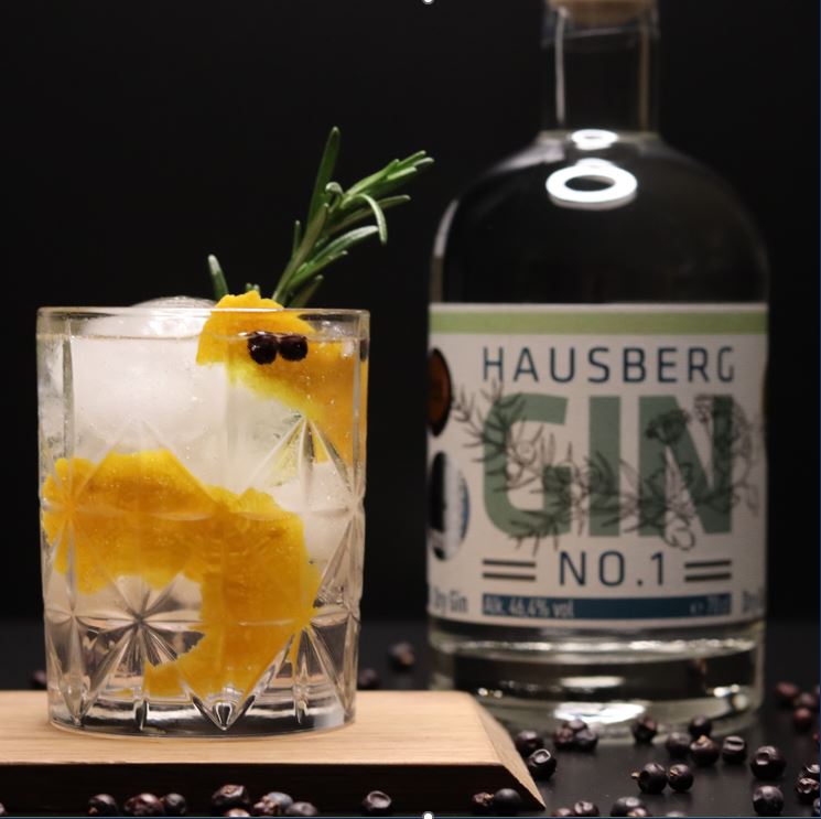 Serviervorschlag für einen Hausberg Gin No.1 im Gin & Tonic. Im Vordergrund steht ein Glas (Tumbler) auf einem Holzbrettchen. Das Glas ist garniert mit Orangenzeste, Rosmarinzweig und zwei Wacholderbeeren.
Im Hintergrund steht eine Flasche Hausberg Gin No.1.
Der Vordergrund ist dekoriert mit verstreutem Wacholder.