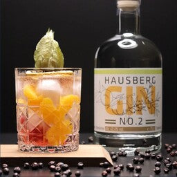 Ein Gin & Tonic im Tumbler (Glas) mit Mandarinen und Beeren als Serviervorschlag im Vordergrund mit einer Flasche Hausberg Gin No.2 rechts seitlich im Hintergrund.
Das Glas steht auf einem Holzbrett. Davor sind Wacholderbeeren dekorativ verstreut, die den Wacholder im Gin symbolisieren. Auf dem Cocktail befindet sich eine Physalis als Garnitur.